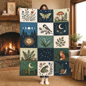 May include: A quilted throw blanket featuring a grid of twelve square panels with nature-themed illustrations including a moth, mushrooms, a bird, a sun, a fox, a frog, a snail, ferns, flowers, a crescent moon, and a star constellation.