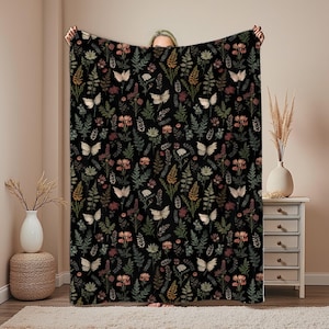 May include: A black blanket featuring a repeating pattern of butterflies, flowers, and foliage in cream, pink, green, and brown. The blanket is held up, displaying the full design. The overall theme is nature and botanical.