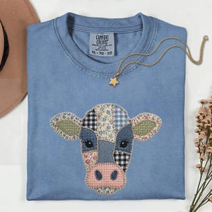 Op de afbeelding: Een lichtblauw Comfort Colors T-shirt met een patchwork koeiengezicht ontwerp. Het gezicht van de koe is gemaakt van verschillende stoffen met patronen, waaronder bloemen, ruitjes en denim. Een gouden sterrenhangerketting ligt op het shirt. Maat XL-TG-EG.