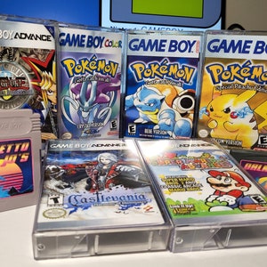 May include: A collection of eight vintage Game Boy and Game Boy Advance video game cartridges. The cartridges are in clear plastic cases and feature various titles, including Pokemon, Yu-Gi-Oh!, Castlevania, and Super Mario Bros. The cartridges are arranged in two rows on a table.