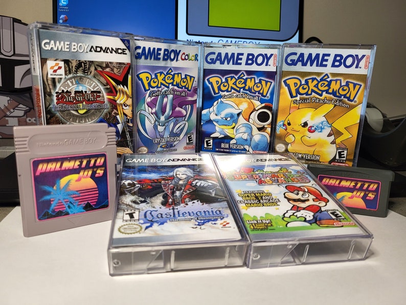 May include: A collection of eight vintage Game Boy and Game Boy Advance video game cartridges. The cartridges are in clear plastic cases and feature various titles, including Pokemon, Yu-Gi-Oh!, Castlevania, and Super Mario Bros. The cartridges are arranged in two rows on a table.