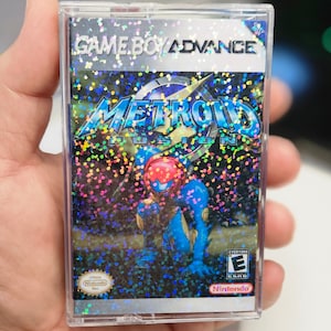 May include: A Game Boy Advance game cartridge with a holographic cover featuring a blue and red character called Metroid Fusion. The game is rated E for Everyone by the ESRB.