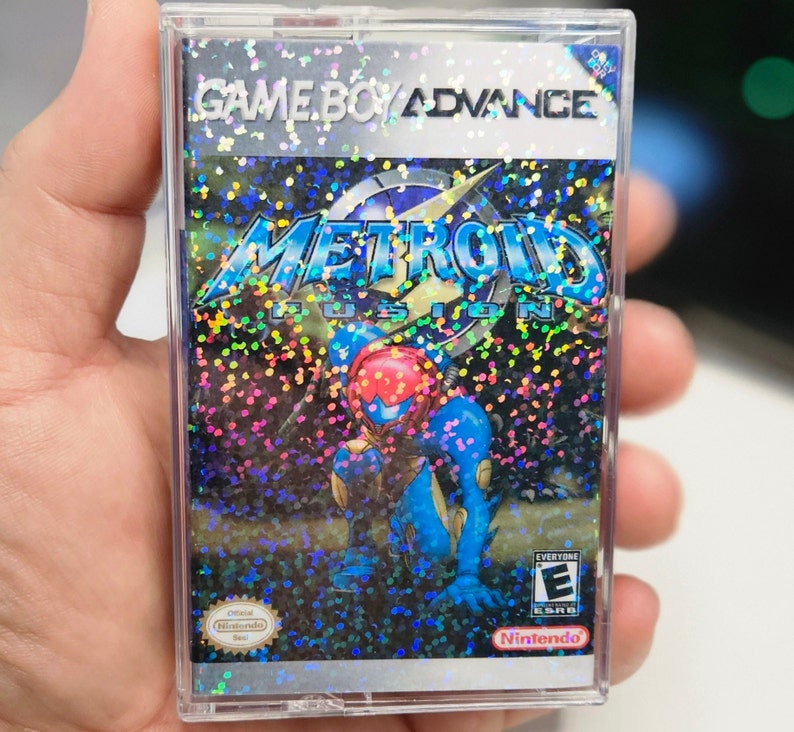 May include: A Game Boy Advance game cartridge with a holographic cover featuring a blue and red character called Metroid Fusion. The game is rated E for Everyone by the ESRB.