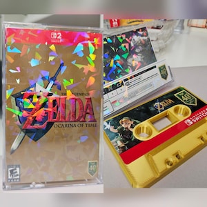 Puede incluir: Una caja de juego de Nintendo Switch para The Legend of Zelda: Ocarina of Time con una cubierta holográfica dorada, junto a un casete de color dorado con una ilustración de Link y el logotipo de Nintendo Switch sobre un fondo verde oscuro.