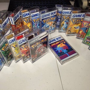 May include: A collection of vintage Game Boy and Game Boy Advance video game cartridges in clear plastic cases. The cartridges feature various Pokemon games, including Ruby, Sapphire, and Emerald versions. There is also a Game Boy cartridge with the text "Palmetto Jr's" on the label.