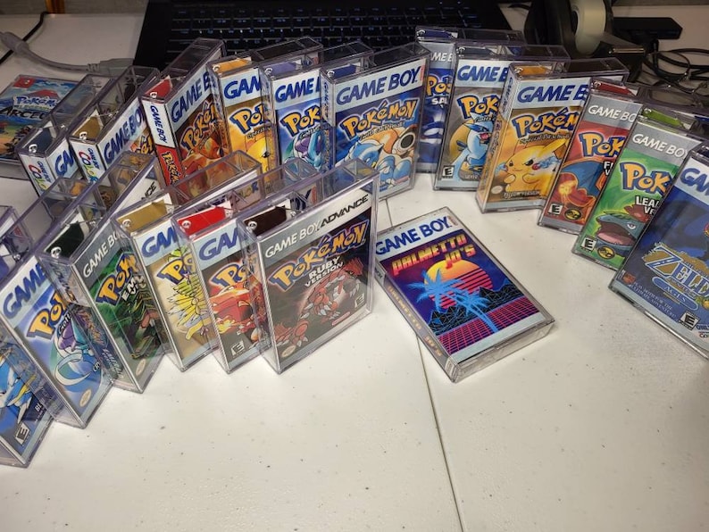 May include: A collection of vintage Game Boy and Game Boy Advance video game cartridges in clear plastic cases. The cartridges feature various Pokemon games, including Ruby, Sapphire, and Emerald versions. There is also a Game Boy cartridge with the text "Palmetto Jr's" on the label.