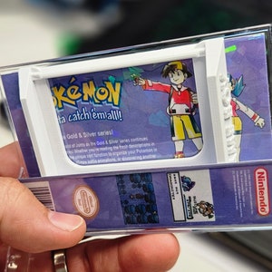 May include: A clear plastic case with a white frame holds a Pokemon video game cartridge. The case has a purple background with the words "Pokemon, Gotta catch 'em all!" and "Gold & Silver series!" printed on it. The image also shows a character from the game, a trainer with red hair and a red shirt, holding a Pokeball.