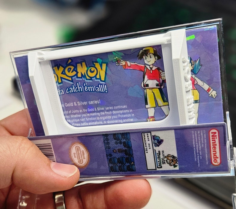 May include: A clear plastic case with a white frame holds a Pokemon video game cartridge. The case has a purple background with the words "Pokemon, Gotta catch 'em all!" and "Gold & Silver series!" printed on it. The image also shows a character from the game, a trainer with red hair and a red shirt, holding a Pokeball.