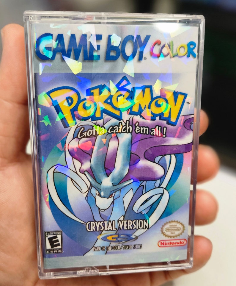 May include: A Game Boy Color game cartridge for Pokemon Crystal Version. The cartridge is silver with a holographic image of a blue and purple Pokemon on the front. The text "Gotta catch 'em all!" is printed on the front of the cartridge.