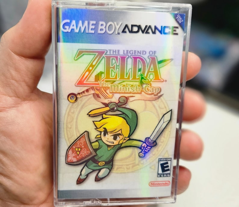 May include: A Game Boy Advance game cartridge for The Legend of Zelda: The Minish Cap. The cartridge is silver with a holographic image of Link, the main character, holding a sword and shield. The game is rated E for Everyone by the ESRB.