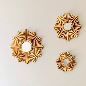 May include: A white desk with gold accents and a laptop, paired with a fluffy white chair. Three gold sunburst mirrors adorn the wall above the desk. The room has a modern, elegant aesthetic.