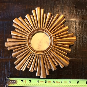 May include: A gold-colored sunburst mirror with a round, reflective center. The mirror's frame features radiating, textured rays. A measuring tape shows the mirror is approximately 11 inches in diameter.
