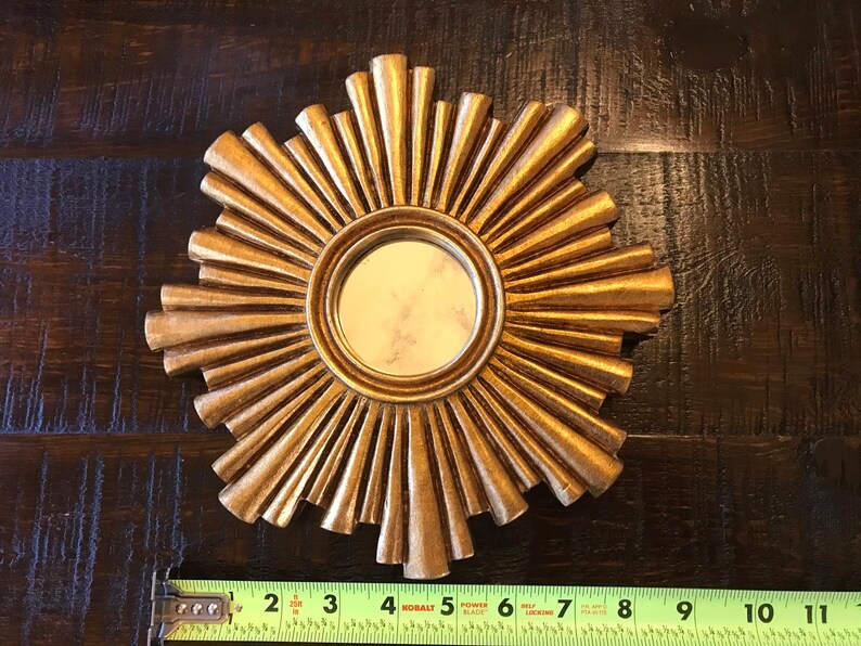May include: A gold-colored sunburst mirror with a round, reflective center. The mirror's frame features radiating, textured rays. A measuring tape shows the mirror is approximately 11 inches in diameter.