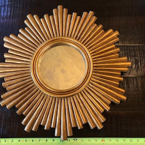 May include: A gold-colored sunburst mirror with a round central mirror. The sunburst design features radiating, textured rays. The mirror measures approximately 11 inches in diameter.