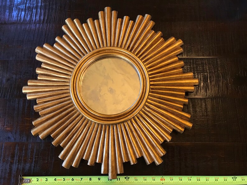 May include: A gold-colored sunburst mirror with a round central mirror. The sunburst design features radiating, textured rays. The mirror measures approximately 11 inches in diameter.