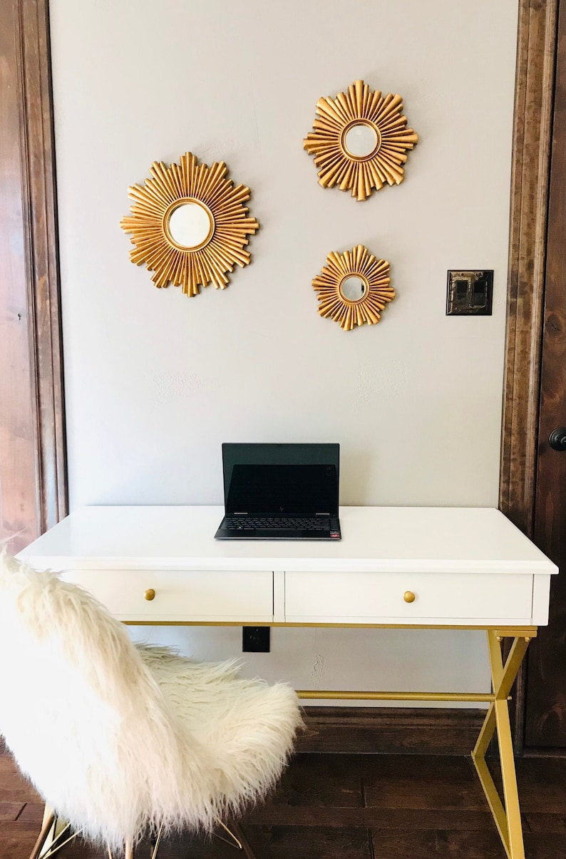 May include: A white desk with gold accents and a laptop, paired with a fluffy white chair. Three gold sunburst mirrors adorn the wall above the desk. The room has a modern, elegant aesthetic.
