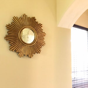 May include: A gold-colored sunburst mirror with a round central mirror. The mirror is mounted on a light beige wall. The sunburst design features radiating, textured rays. The mirror is a decorative wall accent.
