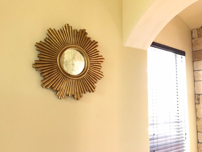 May include: A gold-colored sunburst mirror with a round central mirror. The mirror is mounted on a light beige wall. The sunburst design features radiating, textured rays. The mirror is a decorative wall accent.