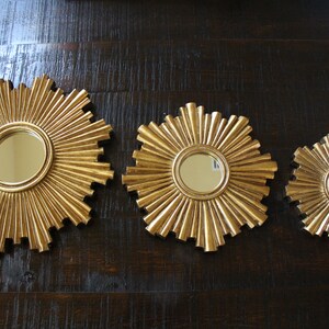 May include: Three gold-colored sunburst mirrors of varying sizes. Each mirror has a circular reflective surface at the center, surrounded by radiating, textured rays. The mirrors are displayed against a dark wood-grain background.