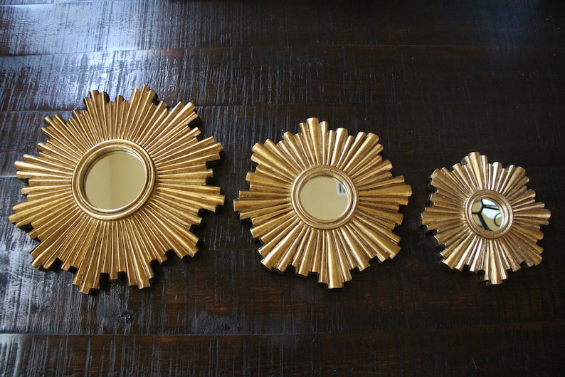 May include: Three gold-colored sunburst mirrors of varying sizes. Each mirror has a circular reflective surface at the center, surrounded by radiating, textured rays. The mirrors are displayed against a dark wood-grain background.