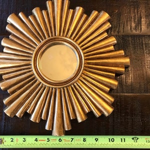 May include: A gold-colored sunburst mirror with a round central mirror. The sunburst design features radiating, textured rays. The mirror measures approximately 11 inches in diameter.