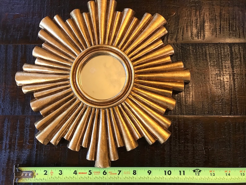 May include: A gold-colored sunburst mirror with a round central mirror. The sunburst design features radiating, textured rays. The mirror measures approximately 11 inches in diameter.