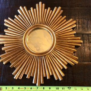 May include: A gold-colored sunburst mirror with a round central mirror. The sunburst design features radiating, pointed elements. The mirror is approximately 12 inches in diameter.