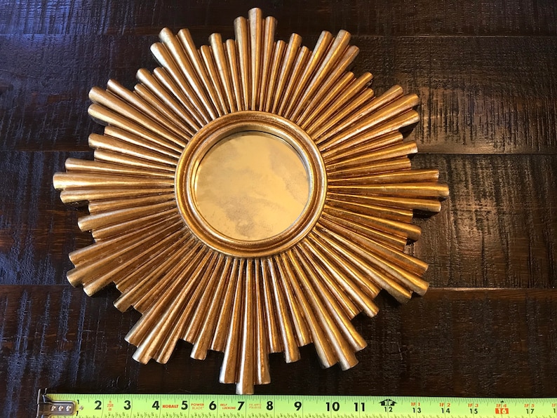 May include: A gold-colored sunburst mirror with a round central mirror. The sunburst design features radiating, pointed elements. The mirror is approximately 12 inches in diameter.