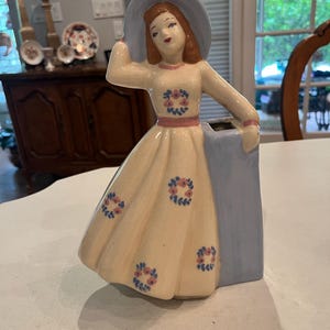 Puede incluir: Figura de cerámica vintage de una mujer con un vestido crema con detalles florales y un sombrero azul. La figura sostiene un objeto rectangular azul, posiblemente un jarrón. El diseño recuerda a la decoración de mediados de siglo.