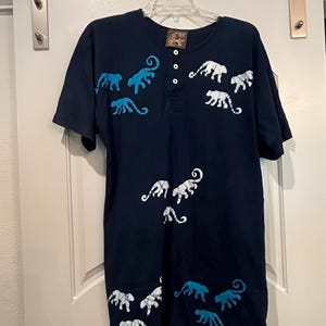 May include: Navy blue short-sleeved shirt with a Henley neckline and a series of buttons. The shirt features a pattern of white and light blue stylized animal figures. The shirt is hanging on a white door.