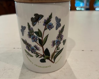May include: A white ceramic jar with a wooden lid. The jar is decorated with a botanical design featuring blue butterflies and flowers. The text "Veronica Chamaedrys" is printed on the jar.