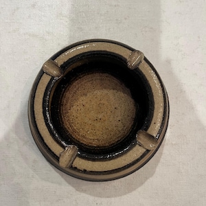 Puede incluir: Un cenicero redondo de cerámica con un diseño de rayas marrones y negras. El cenicero tiene cuatro hendiduras alrededor del borde.