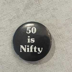 May include: A black button with the text "50 is Nifty" in white. The button is round and has a glossy finish. The text is centered on the button.