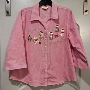 Könnte beinhalten: Ein rosa-weiß gestreiftes Button-Down-Hemd mit Dreiviertelärmeln. Das Hemd zeigt gestickte Katzen mit Weihnachtsmützen und den Text "Santa's Sweeties". Ein festliches Kleidungsstück zum Thema Feiertage.