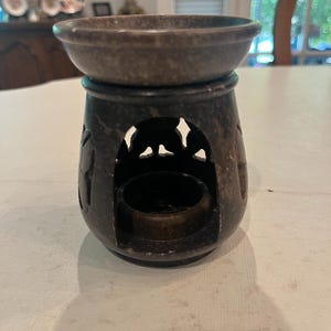 Op de afbeelding: Een bruine stenen etherische olieverspreider met een komvormige bovenkant en een cilindrische basis. De basis heeft decoratieve uitsparingen. De diffuser is ontworpen om een kleine theelichtkaars in de basis en etherische oliën in de bovenste kom te bevatten.