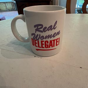 May include: A white ceramic mug with a curved handle featuring the text Real Women DELEGATE! printed on the front. The word Real is in purple, Women is in purple, and DELEGATE! is in red with a red horizontal line underneath the text.