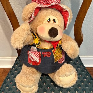 May include: A plush tan teddy bear wearing a red and white gingham dress with a blue denim bib and a red bandana. The bear is sitting on a green chair with a diamond pattern. The bib has the text "Country Bears" embroidered on it.