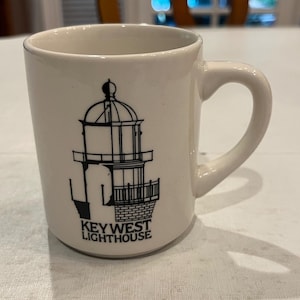 May include: White ceramic mug featuring a black illustration of the Key West Lighthouse. The mug has a curved handle and the words "KEYWEST LIGHTHOUSE" printed below the lighthouse image. The mug is on a white surface.