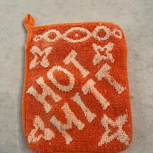 Könnte beinhalten: Orangener Topflappen mit den Worten "HOT SHIT" in weißen Blockbuchstaben. Der Topflappen hat eine Schlaufe zum Aufhängen und dekorative weiße Akzente.