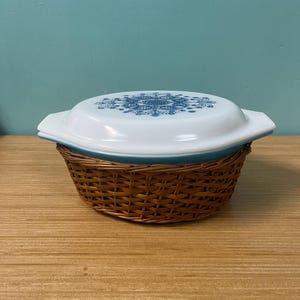 Puede incluir: Una fuente blanca para horno con un diseño floral azul en la tapa, colocada dentro de una cesta tejida marrón. El plato tiene un borde y asas azules. El fondo es de color verde azulado sólido.