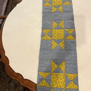 May include: A rectangular quilted table runner featuring three repeating star blocks. Each star is constructed from a textured grey fabric background with bright yellow fabric printed with small red floral patterns forming the star points and center.