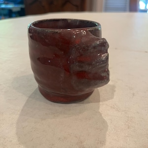 May include: A small, red ceramic mug with a unique, textured handle. The mug has a rustic, handmade look.