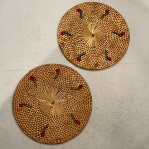 May include: Two round, woven rattan coasters. Each coaster has a central starburst design and is decorated with small red and green floral accents. The coasters are light brown and have a textured surface.