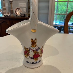 May include: White ceramic basket with a handle, featuring a Mrs. Rabbit illustration with her children. The basket has gold accents on the handle and rim. The base of the basket reads "Mrs. Rabbit".