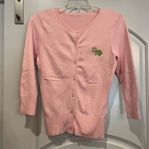 May include: A pale pink cardigan with a V-neck and long sleeves. The sweater features a button-up front and a small embroidered turtle design on the left chest. The garment is suspended from a hanger.