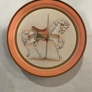 May include: A round, orange-rimmed metal tray featuring a white carousel horse painted on it. The horse has a green and gold saddle and a white mane and tail.