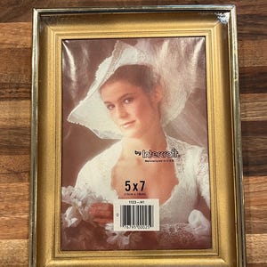 Puede incluir: Un marco de fotos rectangular dorado con una fotografía de una mujer con un vestido de novia. El marco contiene una foto de 13 cm x 18 cm. Fabricado por Intercraft.