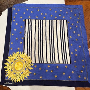 May include: A square silk scarf featuring a central white panel with vertical navy blue stripes, surrounded by a blue border decorated with small yellow stars. A large, detailed yellow sun motif with a face is positioned in the bottom left corner.