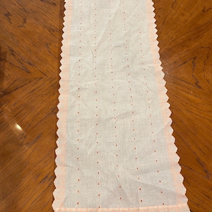 May include: An off-white linen doily with a peach-coloured, scalloped edge. The rectangular cloth has rows of small, evenly spaced, red embroidered dots. The delicate fabric is slightly translucent, suggesting a vintage or handmade item.