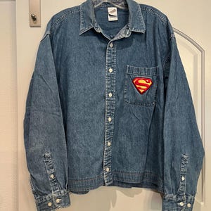 May include: A long-sleeve blue denim button-down shirt featuring a classic collar, a front chest pocket, and a red and yellow Superman logo patch embroidered on the pocket. The shirt has white buttons along the front placket and at the cuffs.